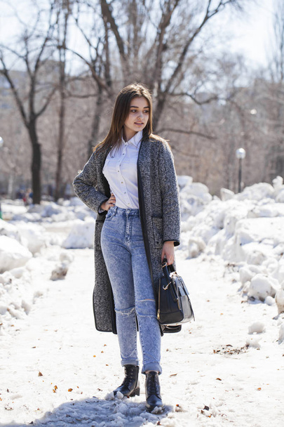 Brunette girl in a gray coat and blue jeans walks in spring park - Photo, image