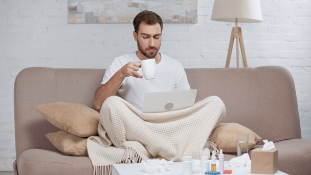 Szakállas blogger ül a kanapén laptop, tüsszörögését, köhögés, figyelembe csésze kávé és ivás a nappaliban  - Felvétel, videó