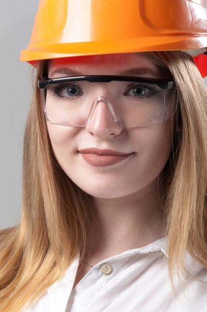 Portrait of a young attractive woman with blond hair in orange helmet on a neutral gray background. - Foto, afbeelding
