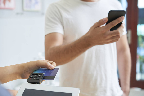 Close-up of man standing with mobile phone while woman making payment by credit card on tablet pc - Foto, imagen