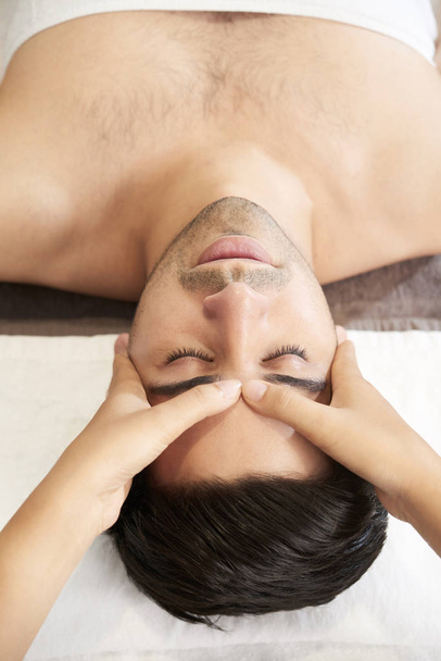 Handsome young man relaxing with eyes closed while therapist massaging his face in spa salon  - 写真・画像