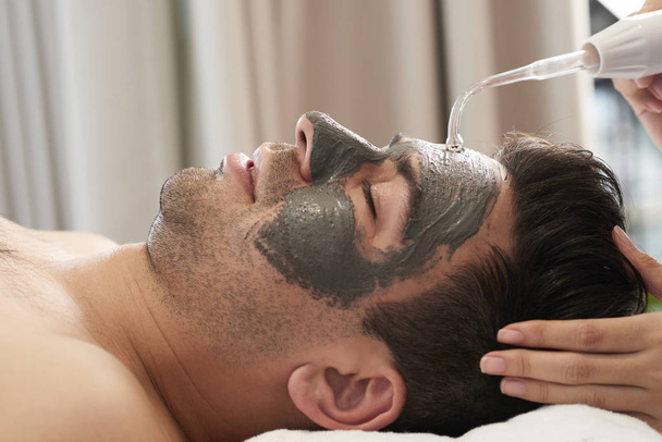 Mladý muž s hliněnou maskou na tváři, který provádí ultrazvukové čištění v lázeňském salonu - Fotografie, Obrázek