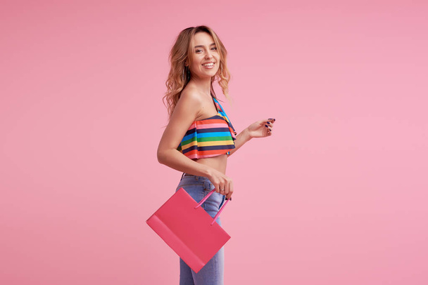 Sale concept. Portrait of an excited beautiful young woman wearing colorful summer clothes holding shopping bag isolated over pink background. Space to sign or text on the package - Valokuva, kuva