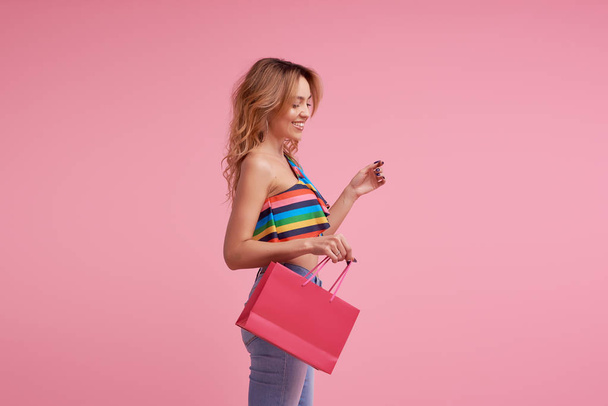 Sale concept. Portrait of an excited beautiful young woman wearing colorful summer clothes holding shopping bag isolated over pink background. Space to sign or text on the package - Fotoğraf, Görsel