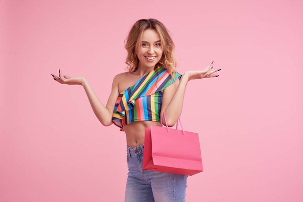Sale concept. Portrait of an excited beautiful young woman wearing colorful summer clothes holding shopping bag isolated over pink background. Space to sign or text on the package - Foto, immagini