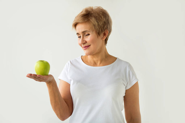 beautiful aged woman with short haircut in a white t-shirt with an apple on a white background - Foto, afbeelding