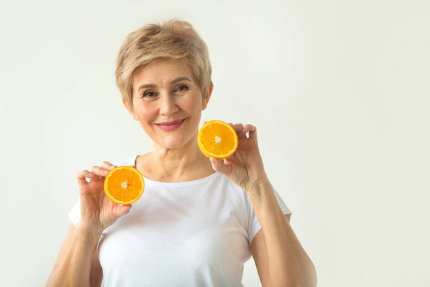 schöne alte Frau mit Kurzhaarschnitt in einem weißen T-Shirt mit Orangen in den Händen auf weißem Hintergrund - Foto, Bild