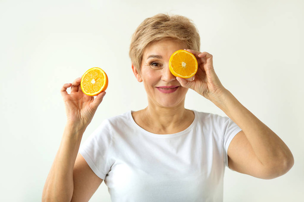 schöne alte Frau mit Kurzhaarschnitt in einem weißen T-Shirt mit Orangen in den Händen auf weißem Hintergrund - Foto, Bild
