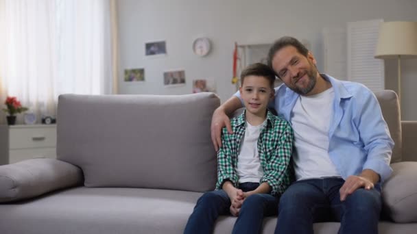 Happy smiling dad and schoolboy son sitting on sofa, looking to camera, family - Video