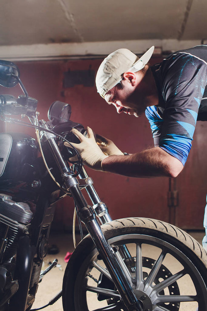 Man Fixing fiets. Zelfverzekerd jonge man repareren motorfiets in de buurt van zijn garage. vervangende lamp in het koplicht - Foto, afbeelding