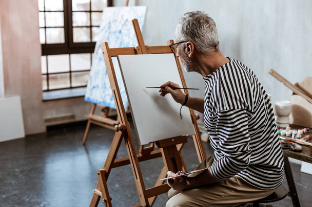 Artist sitting on chair near painting easel while working - Foto, Imagen
