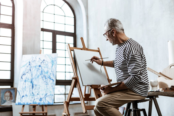 Artist wearing glasses sitting near the window while painting - Foto, imagen