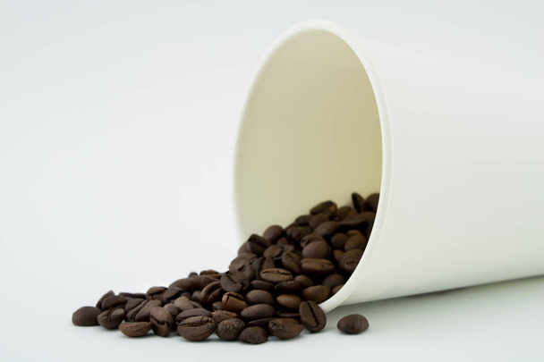 コーヒー豆をこぼしたペーパーコーヒーカップ、白い背景。コーヒー豆. - 写真・画像