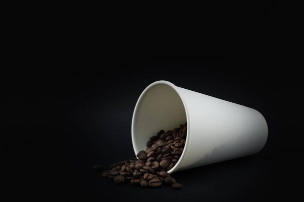 Paper coffee cup with spilled coffee beans, black background. Coffee beans. - Fotó, kép