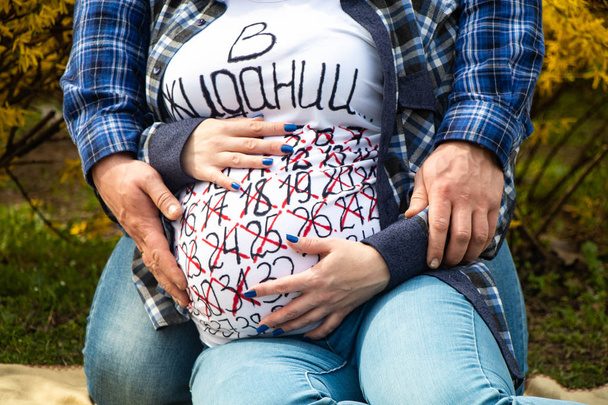Una mujer embarazada vientre se envuelve alrededor de sus brazos maridos, un calendario de embarazo está escrito en una camiseta y las semanas que ya han pasado están tachadas
 - Foto, imagen