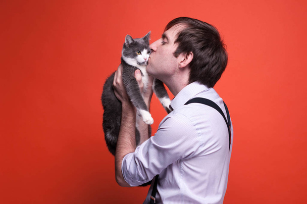 side view of handsome man in shirt with rolled up sleeves and suspender holding and kissing on muzzle grey and white cat on orange background with copy space - Photo, image