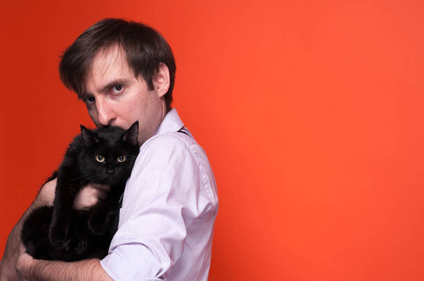side view of handsome serious man, looking at camera, holding and kissing black cat on orange background with copy space - Foto, immagini