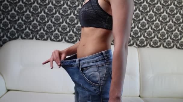 Молодая женщина показывает свою потерю веса и носить ее старые джинсы дома в светлой спальне. Стройная девушка в слишком больших джинсах, показывающая, как она похудела, когда начала есть здоровую пищу
. - Кадры, видео