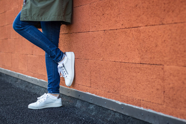 Vrouw in casual kleding en witte sneakers leunend op de muur in de stad. Mode en trendy concept. Vrouwelijke benen met sportschoenen. Kopieerruimte - Foto, afbeelding