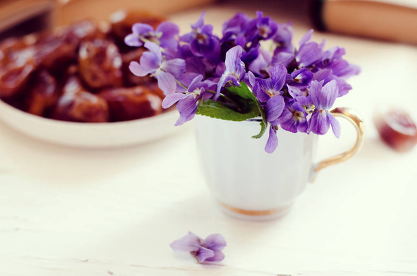 Bodegón con violeta en copa blanca, libros antiguos y frutas de la fecha en un plato. Fondo floral romántico
. - Foto, Imagen