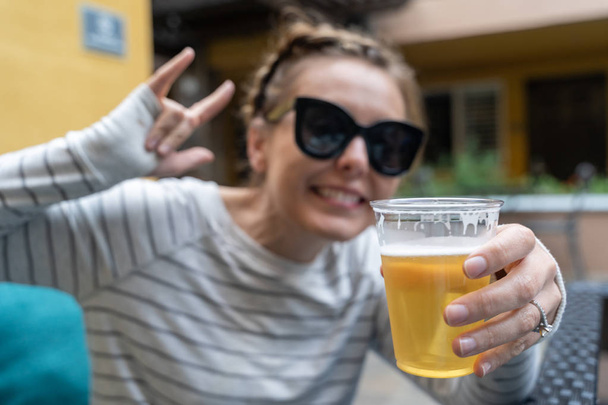 Young woman holds up beer in a plastic cup. Focus on beer, inten - Photo, Image