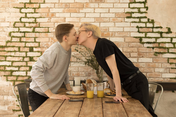 Casal romântico romântico internacional do mesmo sexo beijar uns aos outros em toda a mesa em casa
 - Foto, Imagem