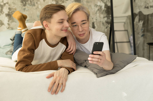 Привлекательная международная гомосексуальная пара отдыхает в постели перед сном и смотрит на экран смартфона
 - Фото, изображение