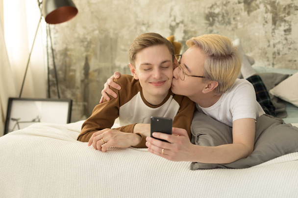 International love. Homosexual asian man with blonde hair kissing his european boyfriend and taking selfie photo - Фото, изображение
