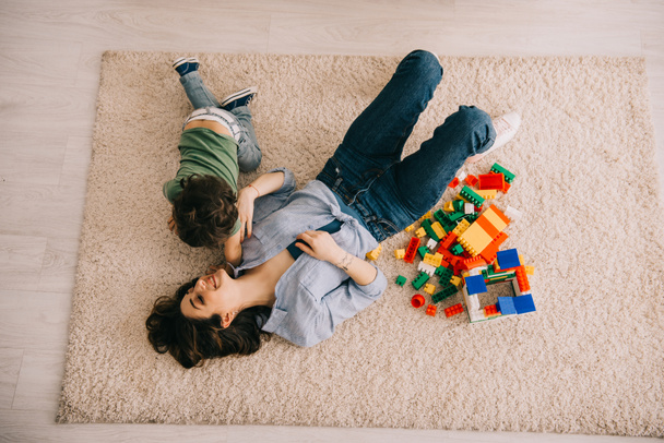 Blick auf lächelnde Mutter und Sohn, die mit Spielzeugklötzen auf Teppich liegen - Foto, Bild