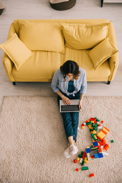 Widok z góry kobieta siedzi na dywan w pobliżu bloków zabawek i za pomocą laptopa - Zdjęcie, obraz