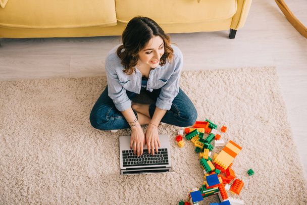 overhead view of smiling woman sitting on carpet near toy blocks and using laptop - Photo, Image