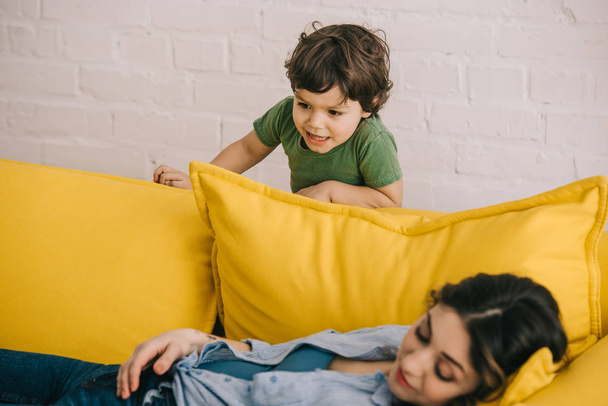 little boy standing near tired mother lying on yellow sofa in living room - Fotó, kép