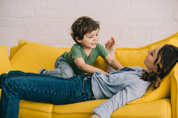 little boy sitting on tired mother while she lying on yellow sofa in living room - Fotografie, Obrázek