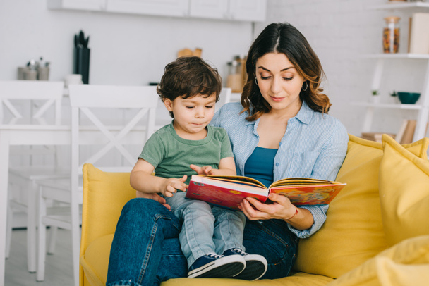 Mom and son sitting on yellow sofa and reading book - Фото, изображение