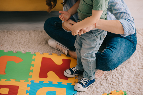 vista parcial de la madre y el hijo con alfabeto alfombra rompecabezas en la alfombra
 - Foto, Imagen