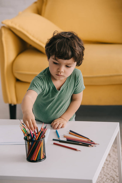 kid in green t-shirt with color pencils and papers in living room - Photo, Image
