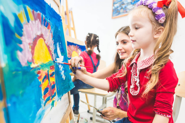 little girl with a teacher in group of preschool student sat the drawing a picture. Painting on maelbert, palette and paints. Back to school and Education concept. - 写真・画像