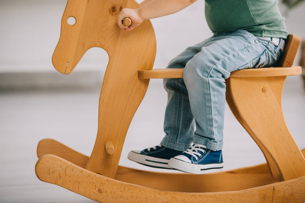 cropped view of child sitting on wooden rocking horse in living room - Fotografie, Obrázek