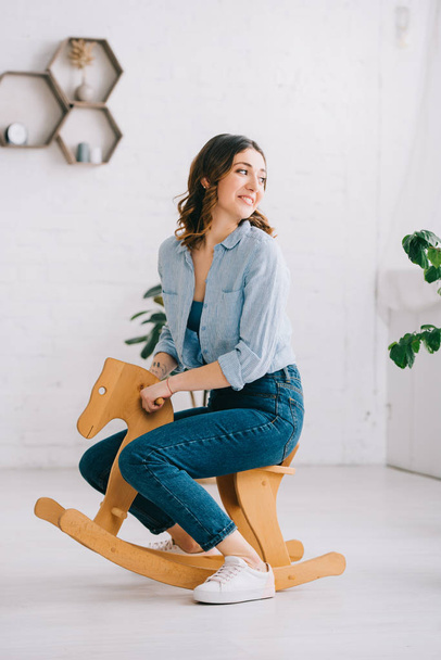 Joyful woman in jeans sitting on rocking horse and looking away - Фото, изображение