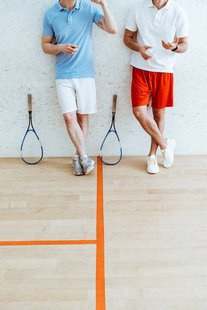 Cropped view of two squash players in shorts standing with crossed legs in four-walled court - Photo, Image