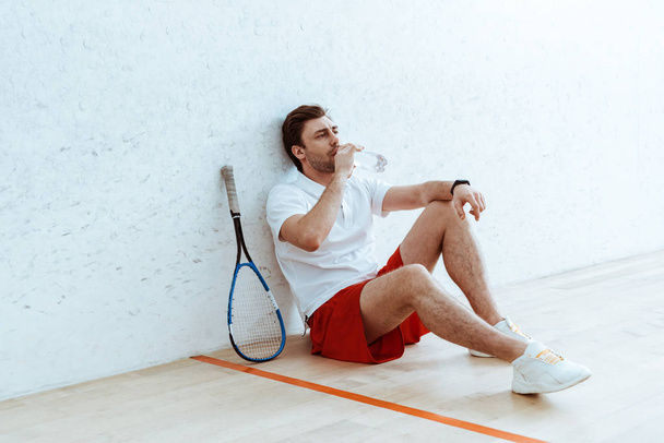 Squash játékos piros nadrágban ülve a padlón és az ivóvízben - Fotó, kép