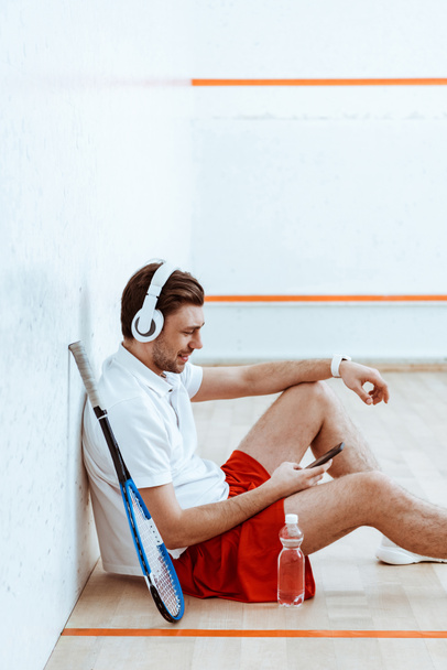 Χαμογελαστός παίκτης σκουός ακούγοντας μουσική στα ακουστικά και χρησιμοποιώντας smartphone - Φωτογραφία, εικόνα