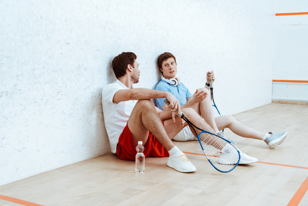 Dwóch graczy squash rozmawiając siedząc na podłodze w czterech ścianach sąd - Zdjęcie, obraz