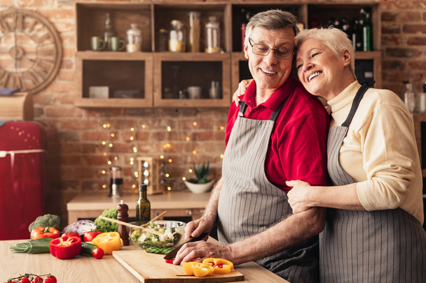 Старша жінка обіймає свого кулінарного чоловіка на кухні
 - Фото, зображення