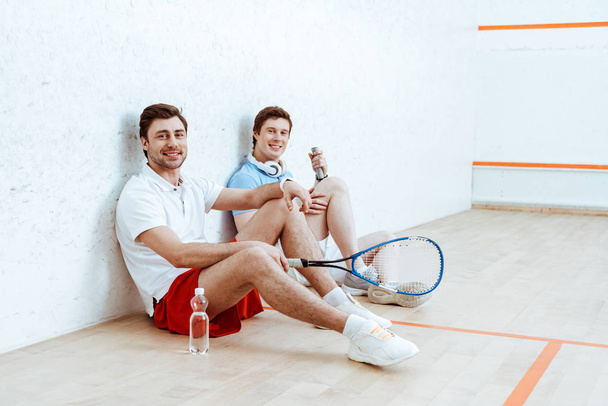 Two smiling squash players sitting on floor in four-walled court - Photo, Image