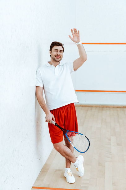 Pełny widok długości odtwarzacza squash w czerwone szorty trzymając rakieta i machając ręką - Zdjęcie, obraz