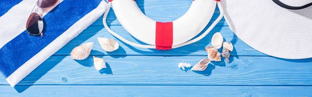 panoramic shot of striped towel, sunglasses, lifebuoy, white floppy hat and seashells on blue wooden background - Photo, Image
