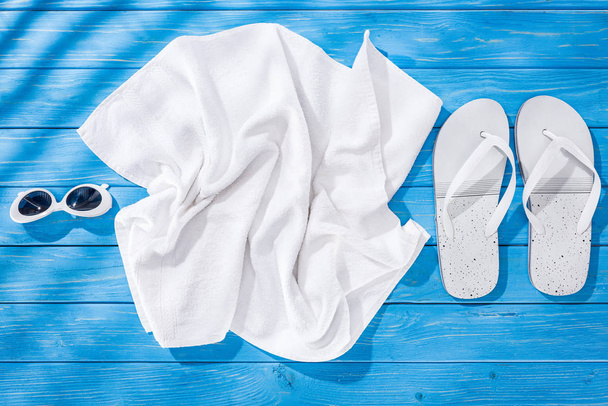 Draufsicht auf weißes zerknülltes Handtuch, Sonnenbrille und Flip-Flops auf blauem Holzhintergrund - Foto, Bild