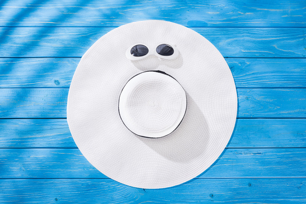 верхний вид белых солнцезащитных очков и белая гибкая шляпа с черной лентой на синем деревянном фоне
 - Фото, изображение