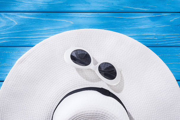 top view of white sunglasses and white floppy hat with black ribbon on blue wooden background - Fotó, kép
